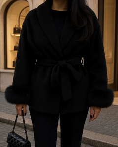 Elegante schwarze Damenjacke mit Bindegürtel und Kunstfell-Besatz an den Ärmeln, kombiniert mit schwarzer Hose und Designerhandtasche vor einer Luxus-Boutique in Mailand.