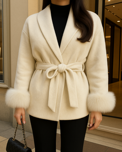 Elegante Frau trägt eine cremefarbene Wolljacke mit Bindegürtel und Fellmanschetten, kombiniert mit schwarzer Hose und Chanel-Handtasche, vor einer modernen Boutique.