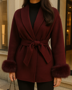 Frau trägt einen eleganten bordeauxfarbenen Wollmantel mit Bindegürtel und Fellmanschetten, kombiniert mit schwarzer Hose, vor einer modernen Boutique.