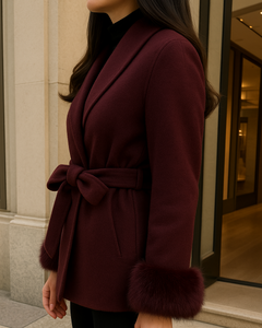 Frau trägt einen eleganten bordeauxfarbenen Wollmantel mit Bindegürtel und Fellmanschetten, kombiniert mit schwarzer Hose, vor einer modernen Boutique.