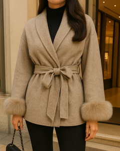 Frau trägt einen eleganten beigefarbenen Wollmantel mit Gürtel und Fellmanschetten, kombiniert mit schwarzer Hose und Chanel-Handtasche, vor exklusiver Boutique.