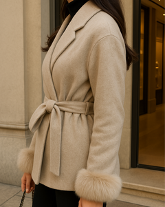 Frau trägt einen eleganten beigefarbenen Wollmantel mit Gürtel und Fellmanschetten, kombiniert mit schwarzer Hose und Chanel-Handtasche, vor exklusiver Boutique.